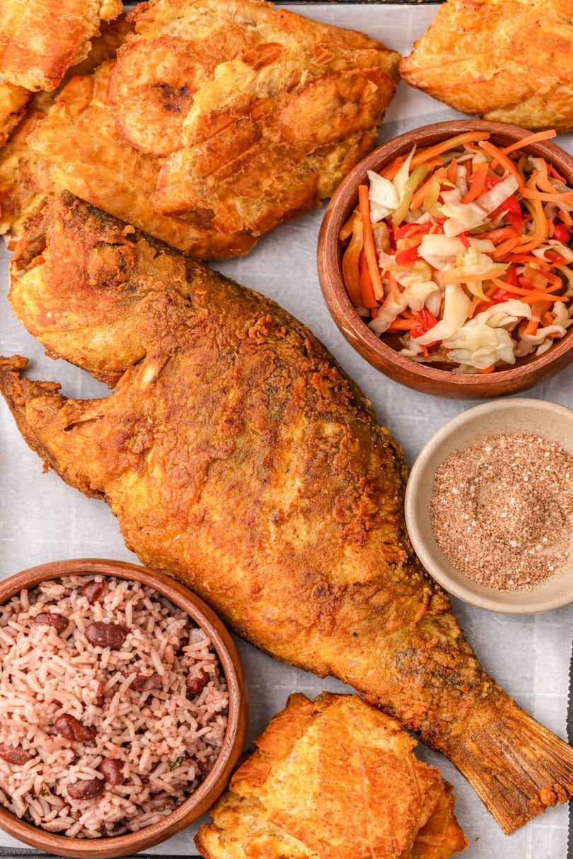 Haitian fried fish, rice and beans, pikliz, and fried plantains on a white surface.