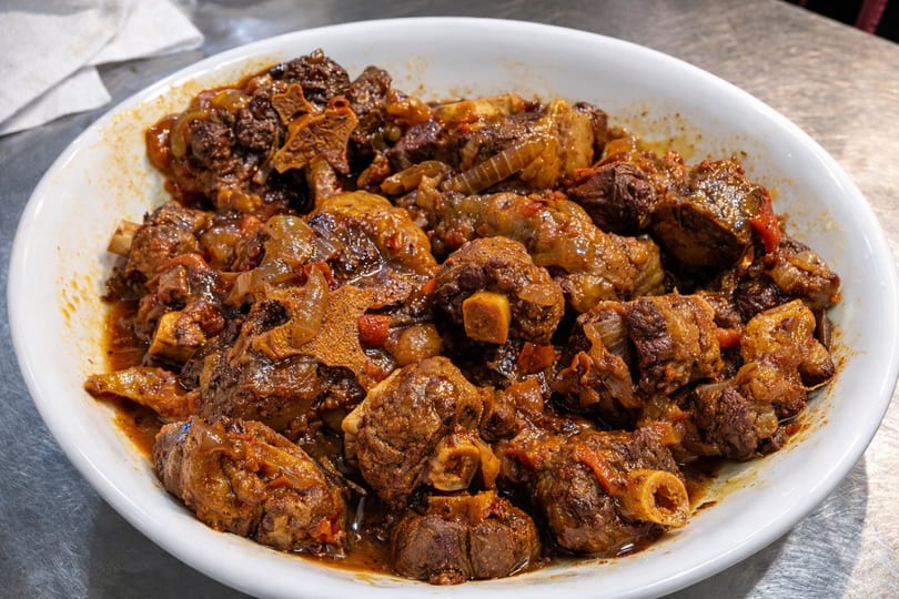 A white bowl filled with rich, brown Haitian oxtail stew, featuring meat, onions, and peppers.